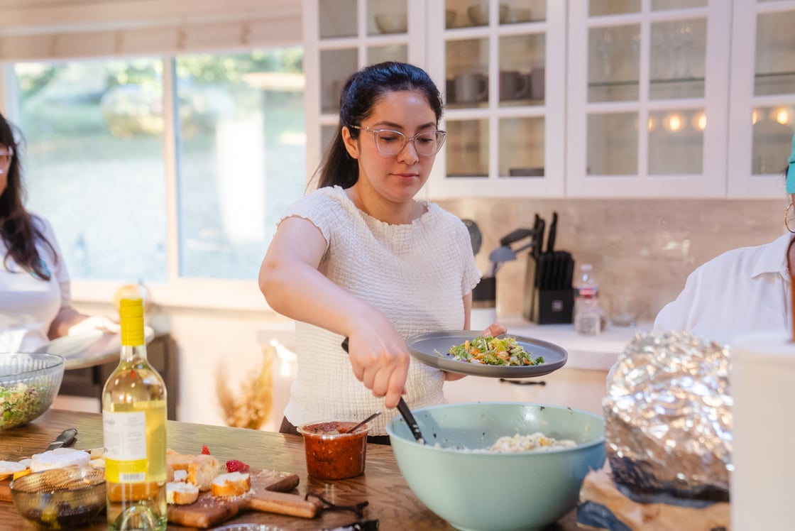 A woman in a white shirt with glasses and brown hair scoops salad out of a large bowl next to many more food items.