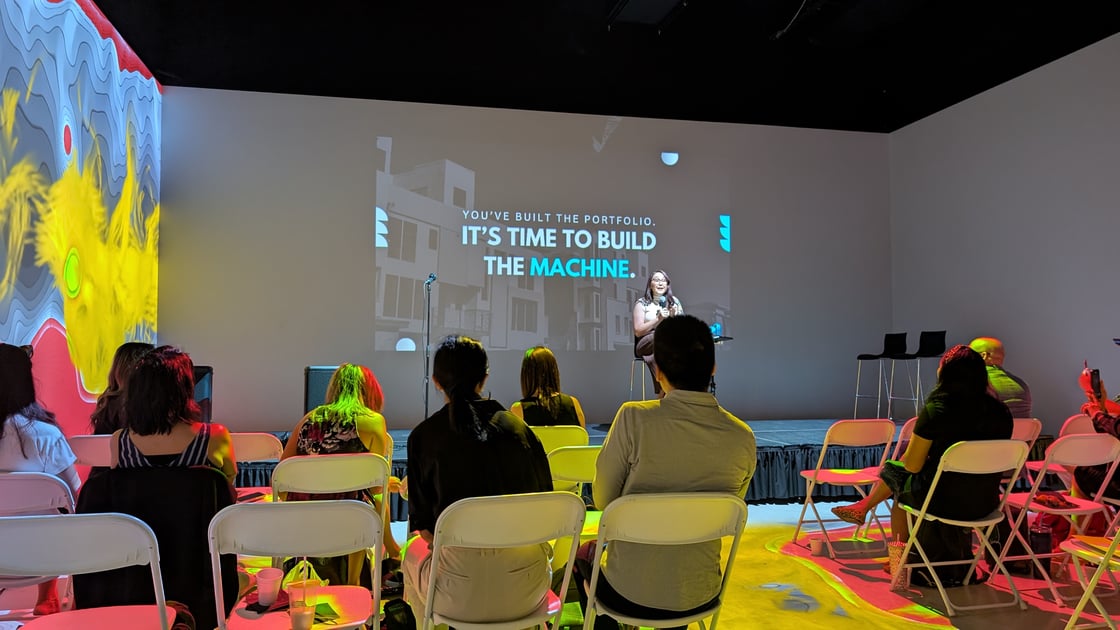 A woman sits on stage speaking to an audience of people in a dark room lit with projection art. Behind the speaker is a presentation with the words "You've built the portfolio, It's time to build the machine."